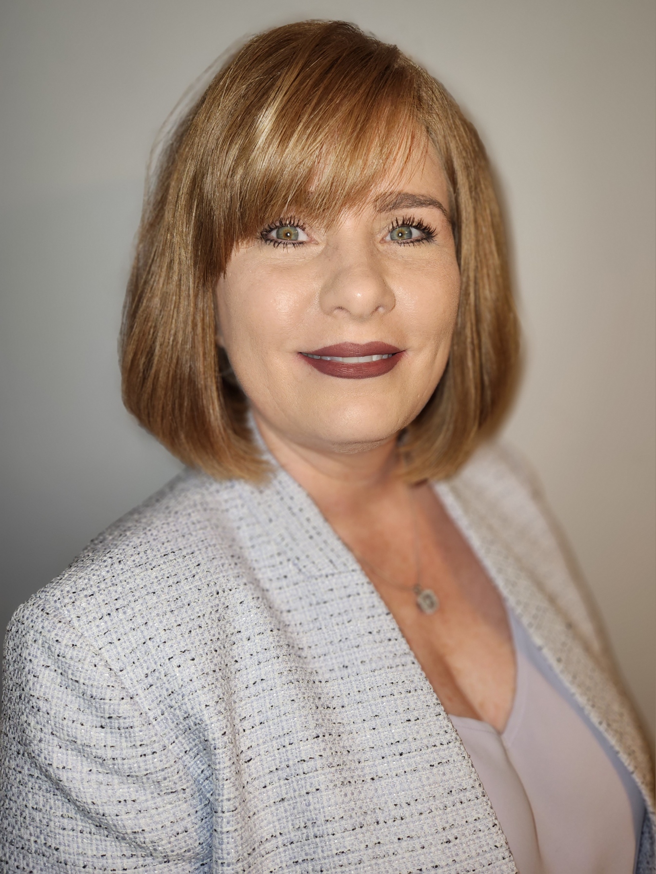 Woman with auburn bob haircut and bangs wearing light gray textured blazer, smiling at camera against gray background